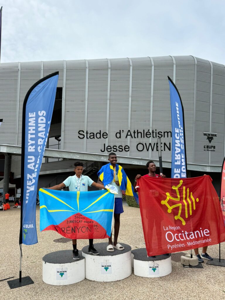 3 jeunes sur un podium tenant des drapeaux car ils ont gagnés le championnat de france de para athlétisme. 1 de ces jeunes fait partie de l'IME les Capitelles de l'Apeai Ouest Hérault